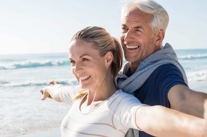 Happy senior couple standing on beach with arms outstretched and looking away. Happy couple at beach on a bright sunny day. Retired husband and smiling wife thinking about their future.